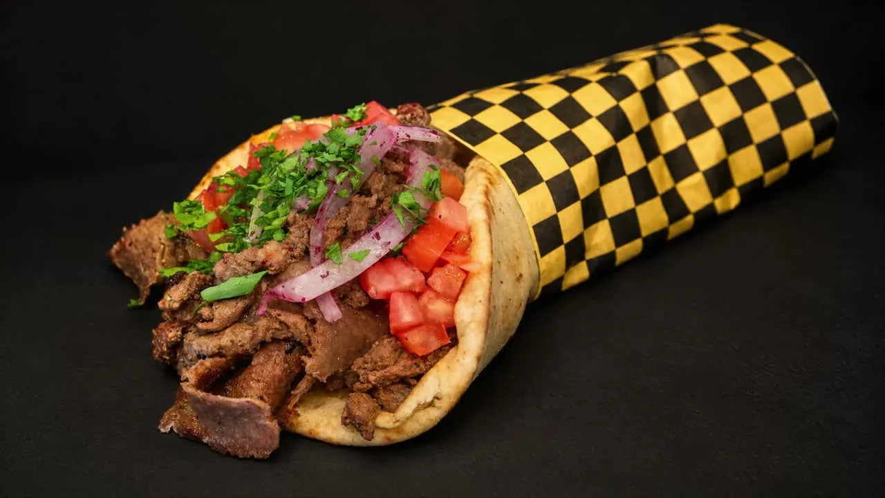 A gyro pita wrapped in black and yellow checkered paper, piled with shaved gyro, tomato, red onion, parsley and tzatziki