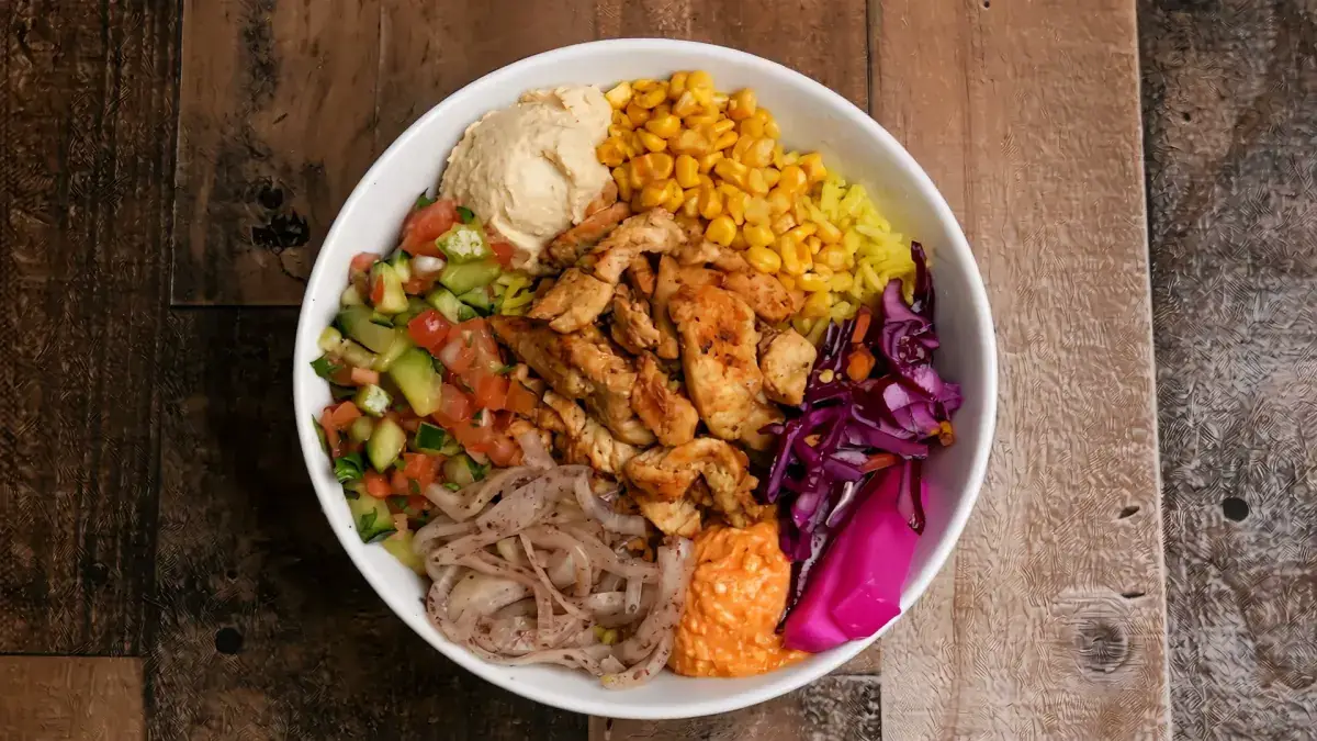 A bowl filled with hummus, corn, cabbage, pickled turnips and tomato cucumber salad